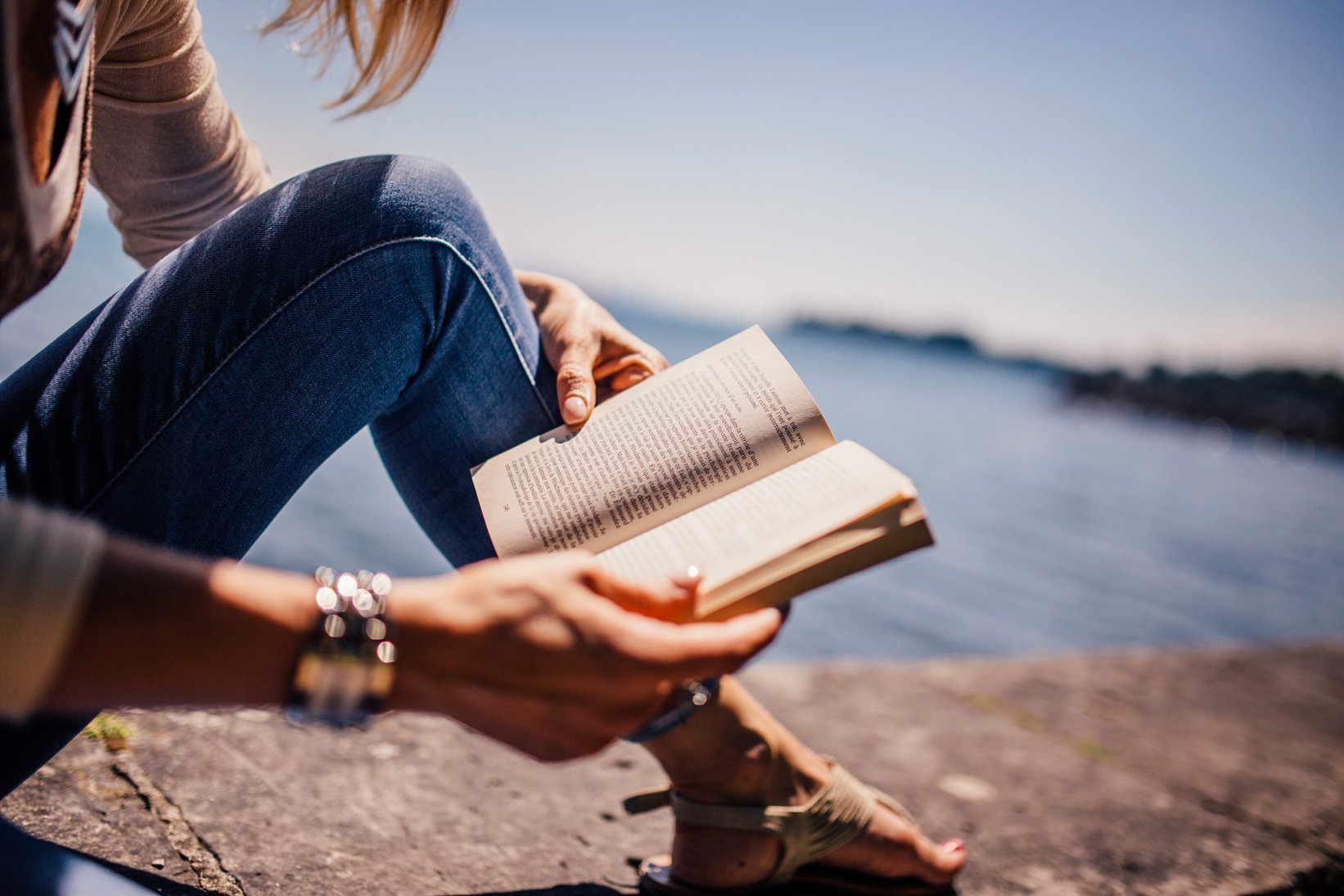 Woman Wearing Blue Denim Jeans Holding Book Sitting on Gray Concrete at Daytime